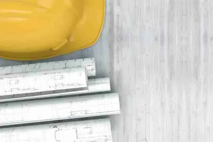 Yellow hard hat resting on rolled blueprints on a whitewashed wood table