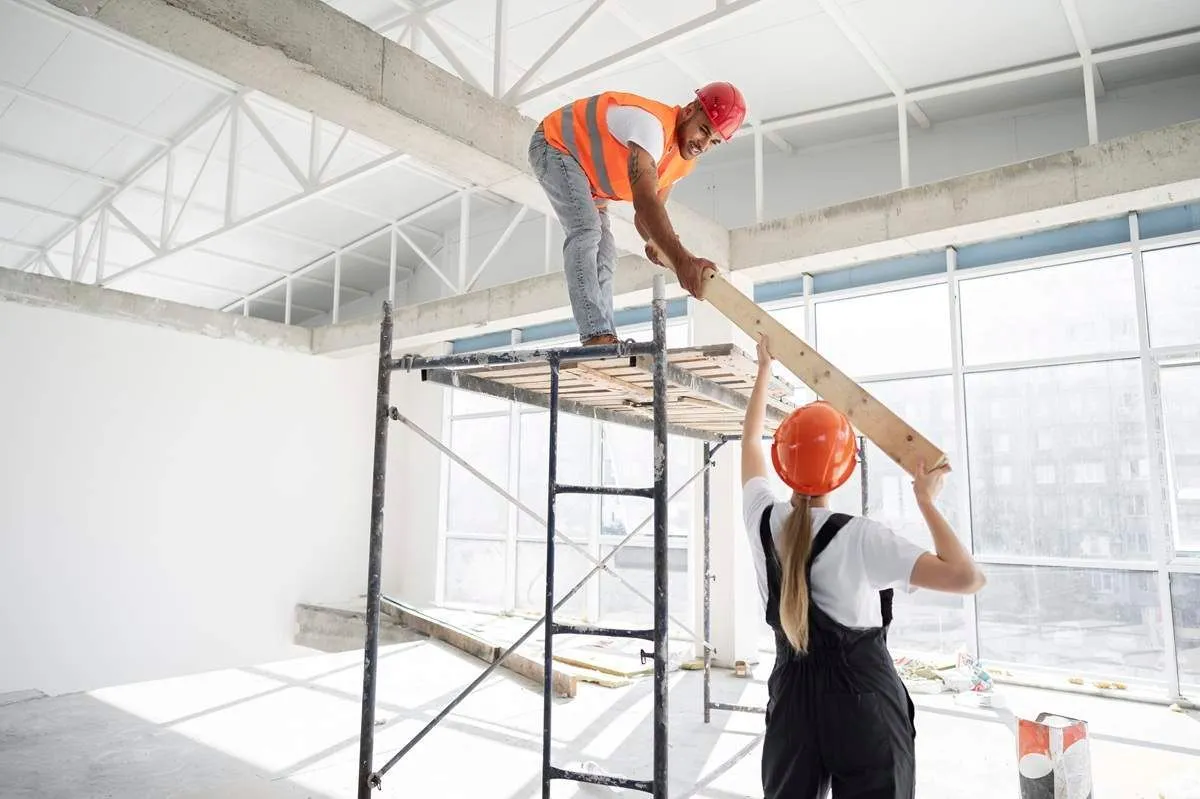Two workers on scaffolding passing a steel beam inside an unfinished commercial space