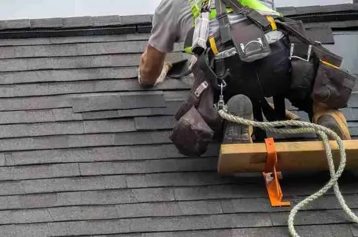 Roofer in a safety harness installing asphalt shingles on a steep roof